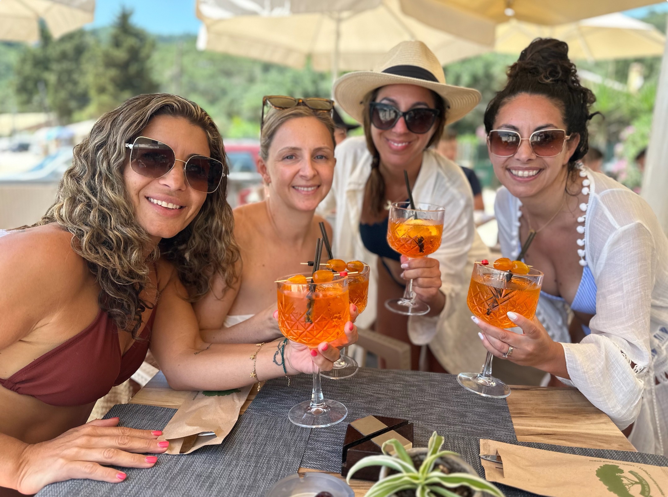 Four guests smiling and raising spritz glasses under beach umbrellas